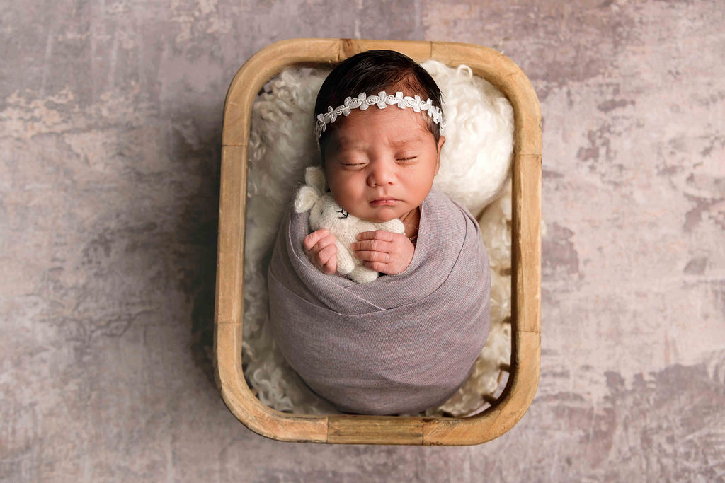 A newborn baby sleeps in a swaddle holding a teddy in a wooden basket after visiting little talkers in stamford, ct
