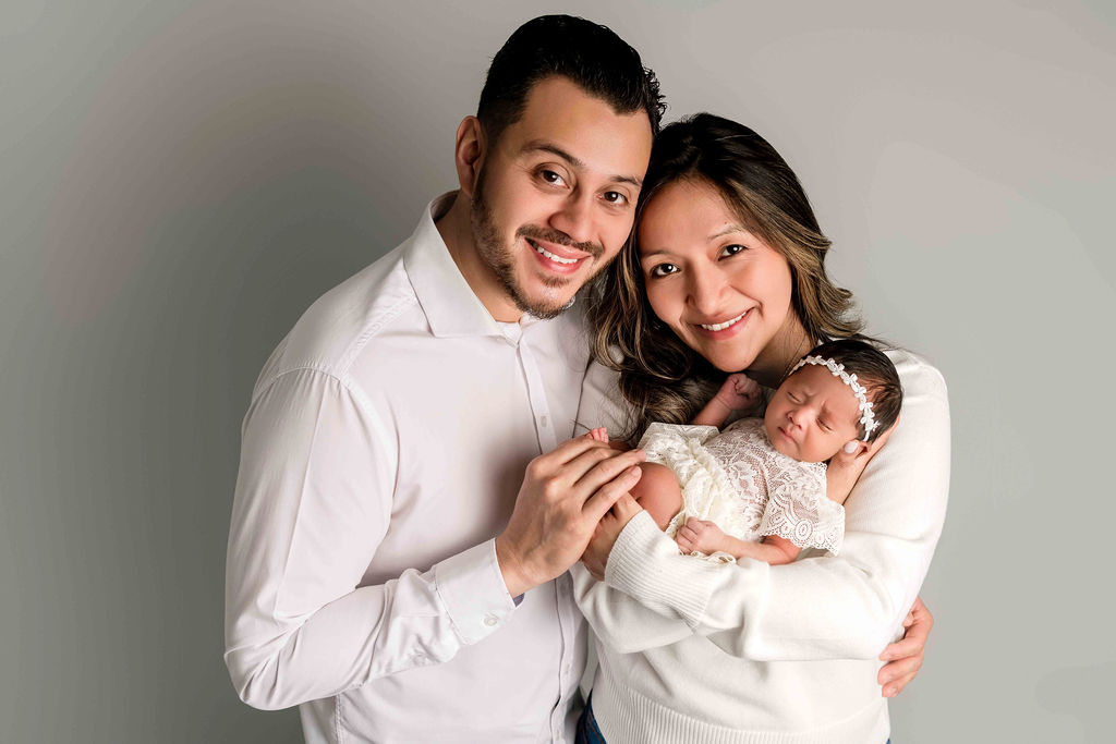 Happy new mom and dad in white snuggle their sleeping newborn against mom's chest in a studio after visiting little talkers in stamford, ct