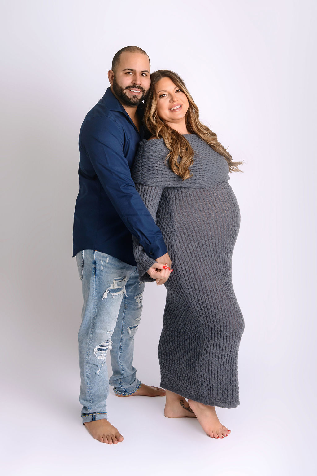 Happy expecting parents in blue snuggle and hold hands while standing in a studio