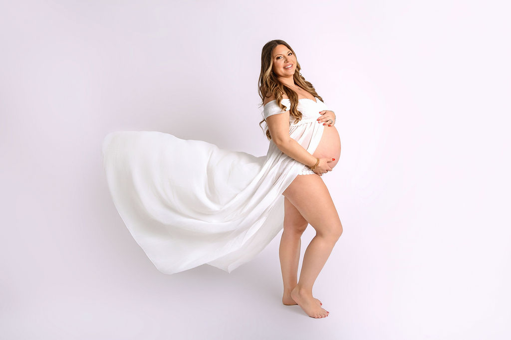 A smiling mother to be stands holding her bump in a studio while wearing an open front white maternity dress after meeting with midwives in Stamford, CT