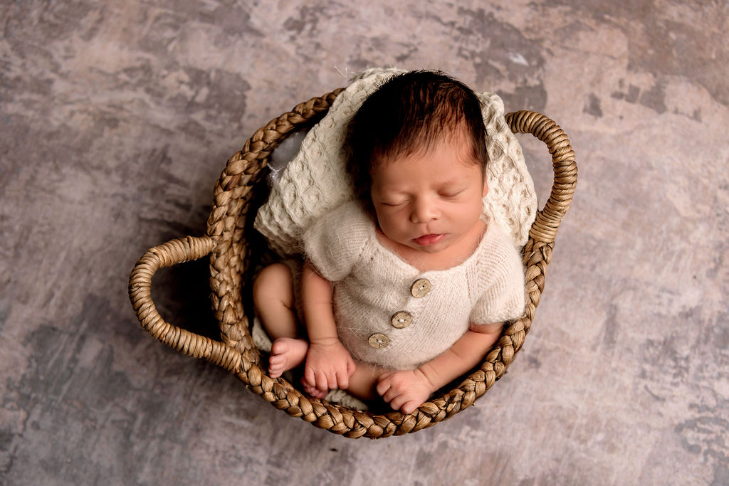 A sleeping newborn sits in a woven basket wearing a white knit onesie after visiting Wee Mondine