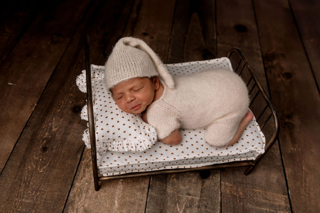 A newborn baby sleeps on a tiny metal bed in a white knit onesie and matching sleep cap after visiting Wee Mondine