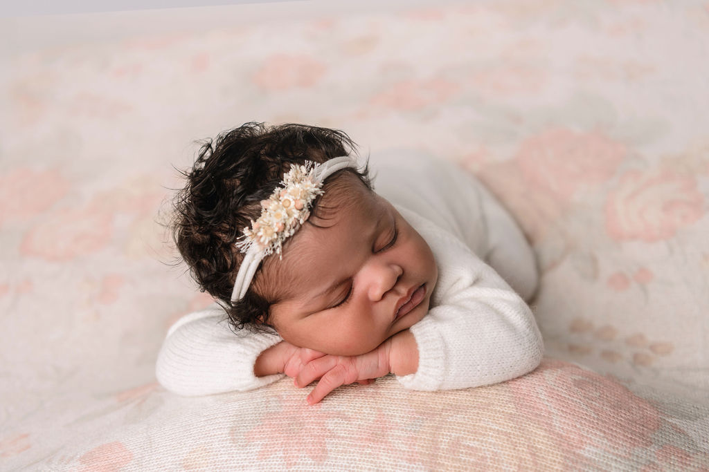 A newborn baby sleeping with cheek on her hands on a flower print bed in a studio