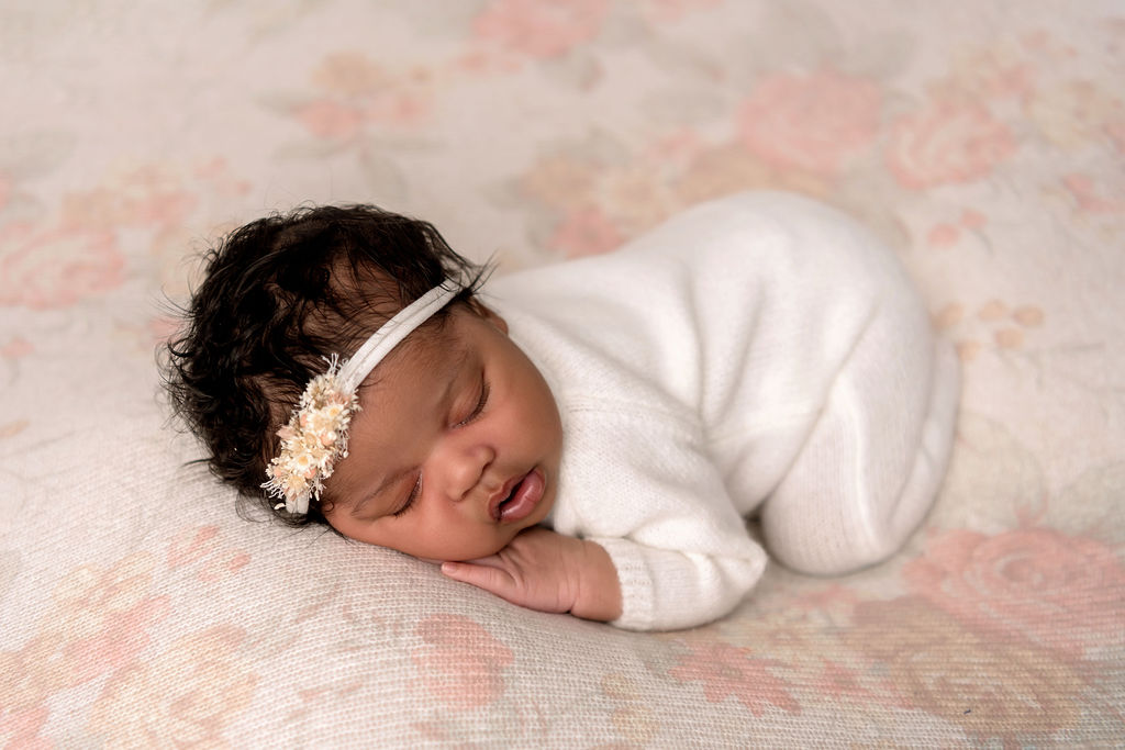 A newborn baby girl on a flower print bed sleeping in a white onesie