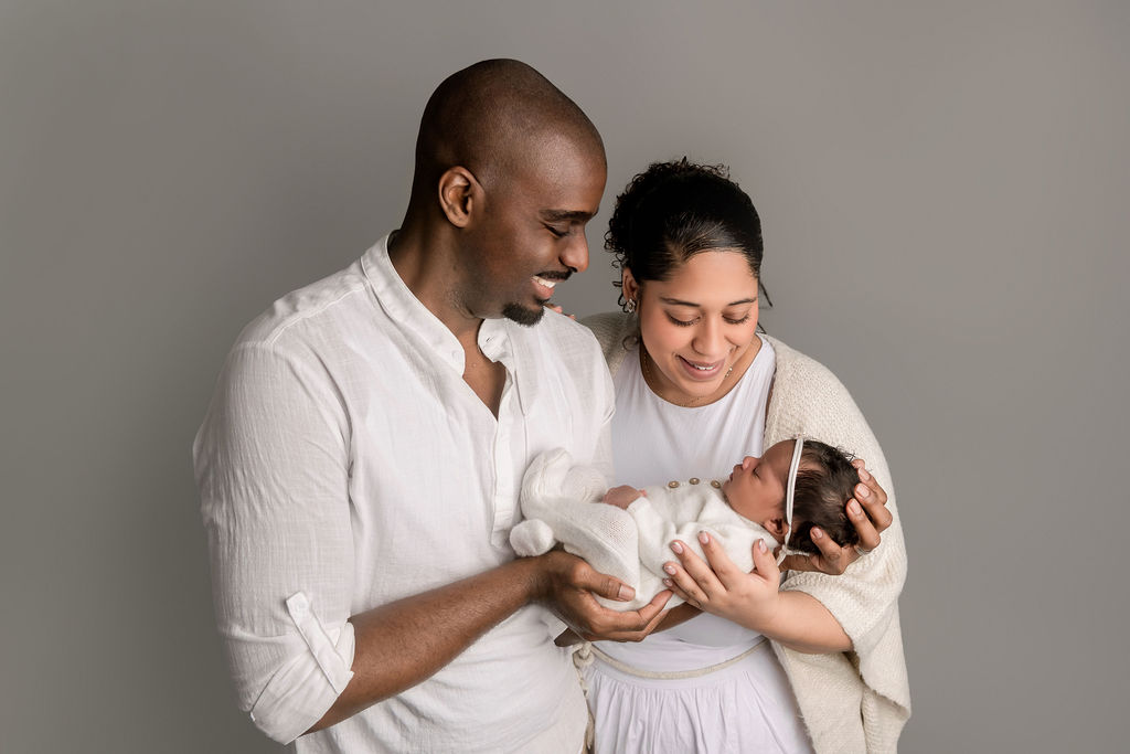 Happy new mom and dad in white smile down to their newborn cradled and sleeping in their hands while standing in a studio thanks to Connecticut Childbirth & Women’s Center