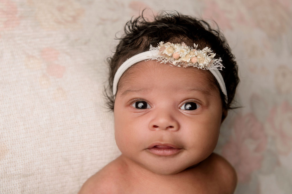 A curious newborn lays with eyes open and a flower headband on a pink flower bed after leaving Connecticut Childbirth & Women’s Center