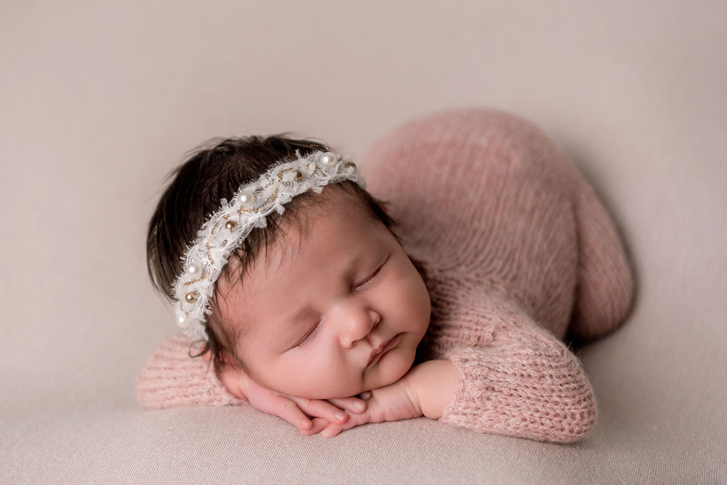 A sleeping newborn baby girl with a pearl headband and pink knit onesie