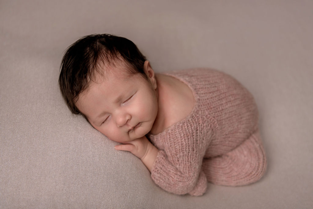 A sleeping newborn baby in froggy pose in a pink knit onesie