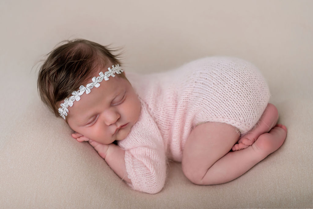 A newborn baby sleeps in froggy pose in a pink knit onesie and tiny headband