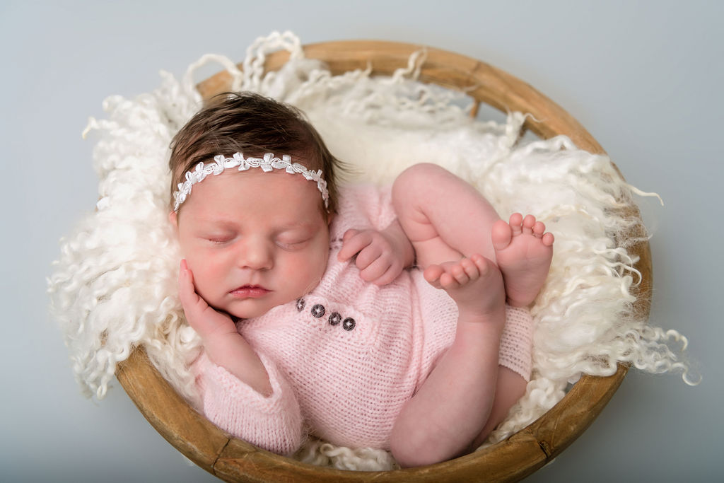 A sleeping newborn baby girl in a knt pink onesie in a wooden bowl and white blanket