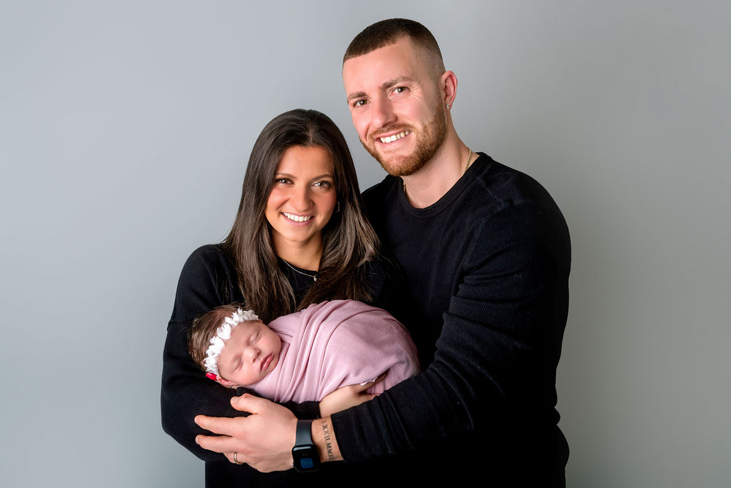 Smiling mom and dad in matching black sweaters stand holding their sleeping newborn after meeting postpartum doulas in Stamford, CT