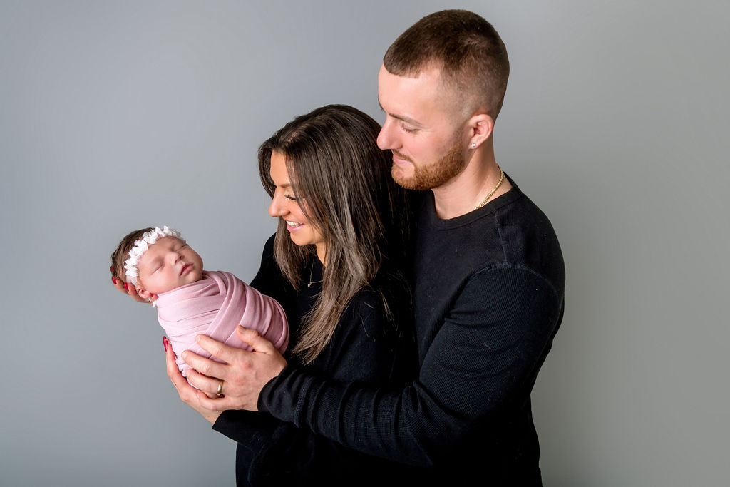 Happy new parents in black smile at their sleeping newborn in their hands in a pink swaddle after meeting postpartum doulas in Stamford, CT