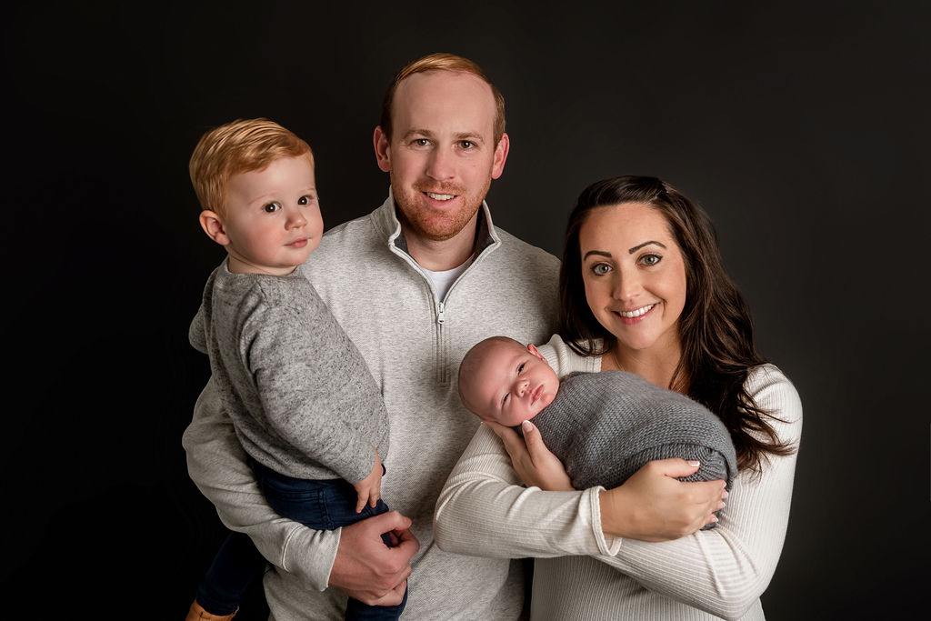 A happy mom and dad stand in a studio in white sweaters holding their newborn and toddler son