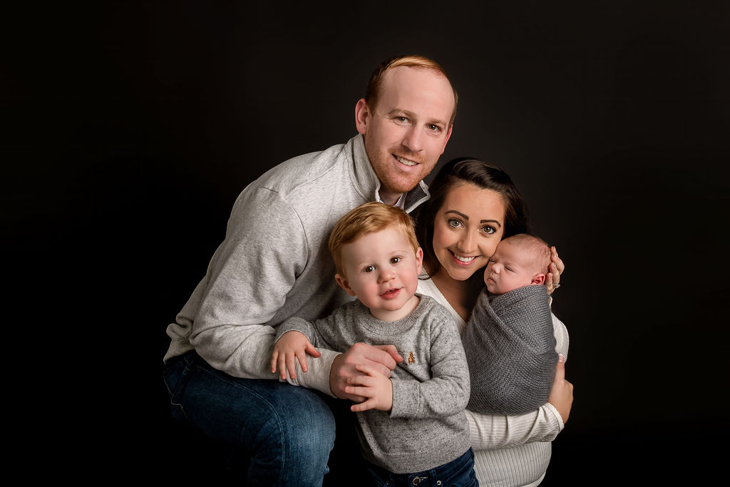 Smiling mom and dad hug and cradle their toddler and newborn in grey