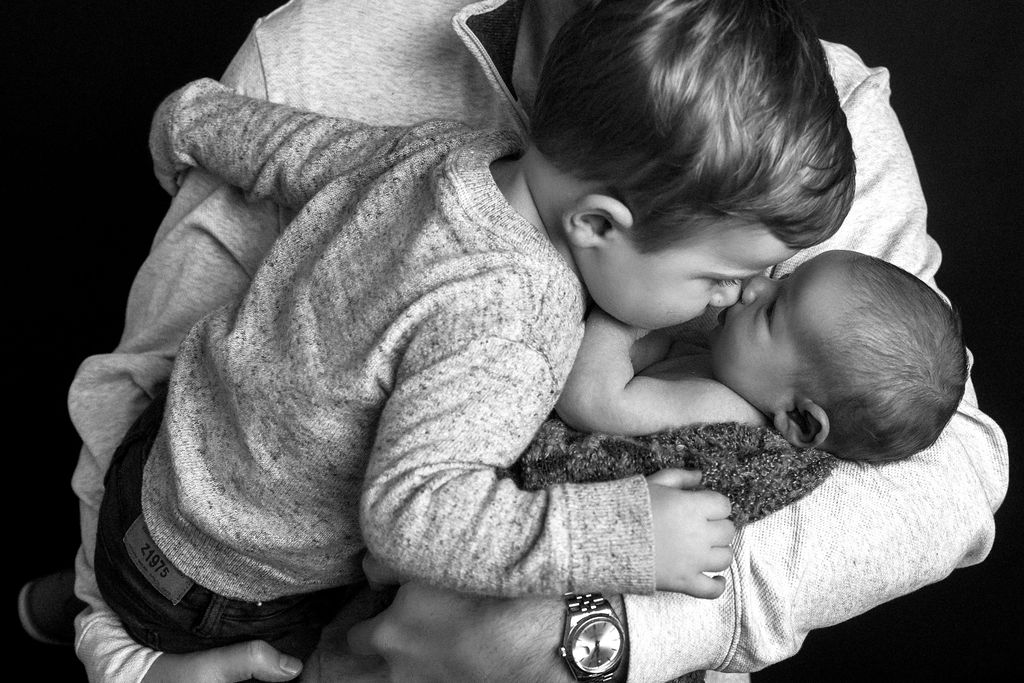 A toddler boy leans over to kiss newborn baby sibling while both in dad's arms in balck and white after mom used prenatal chiropractors in Stamford, CT