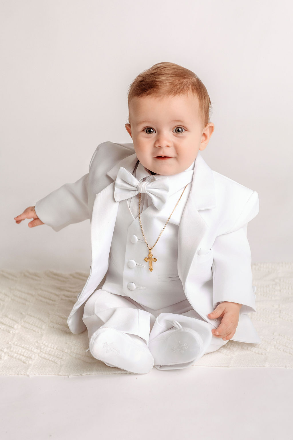 A happy baby in a full white suit and jacket sitting on a rug in a studio