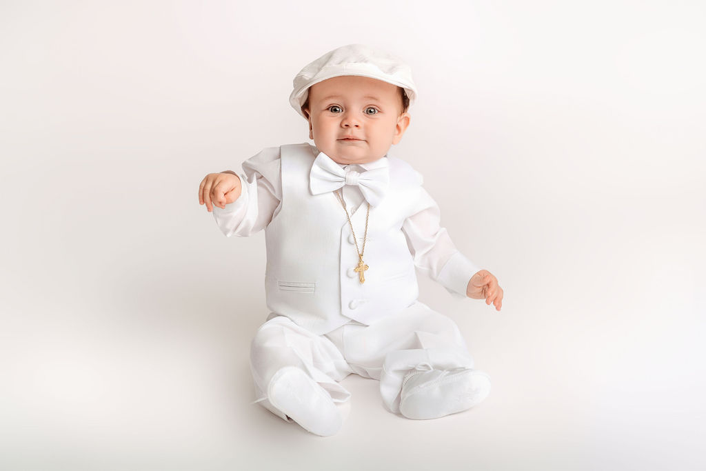 A happy baby in a hat and white suit with gold cross and bowtie sitting in a studio