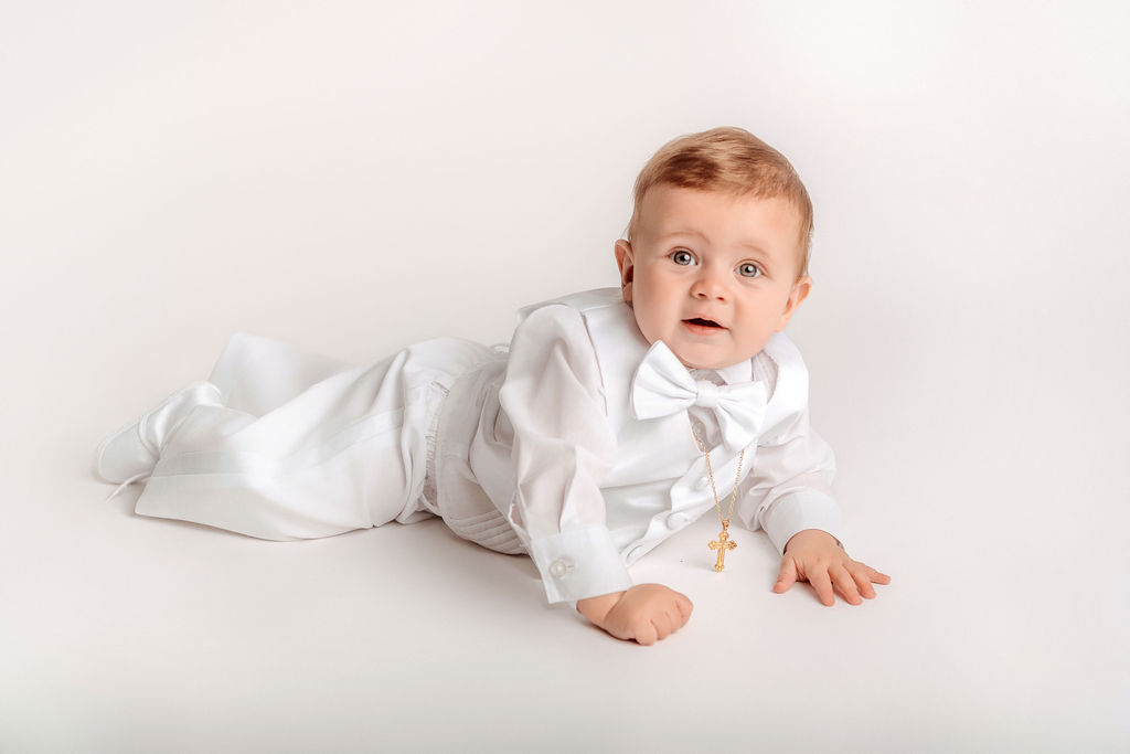 A crawling baby in a white suit and bowtie with a gold cross chain in a studio before visiting children's museums in connecticut