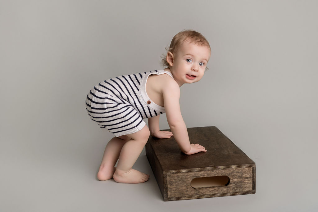 A toddler bends over to climb up an apple box in a studio wearing stripe knit overalls