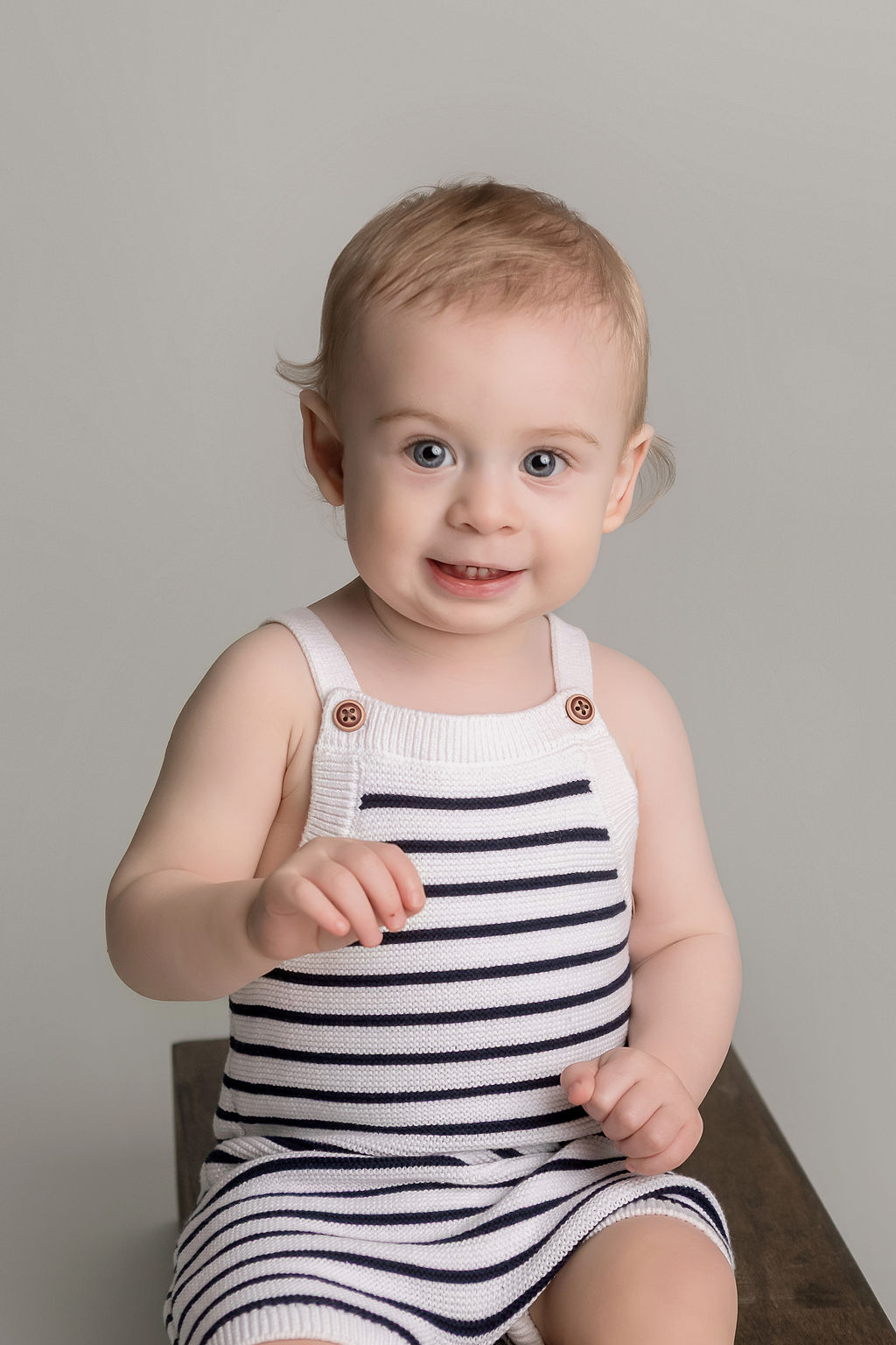 A happy young toddler smiles while sitting in knit stripe overalls on a wooden box in a studio