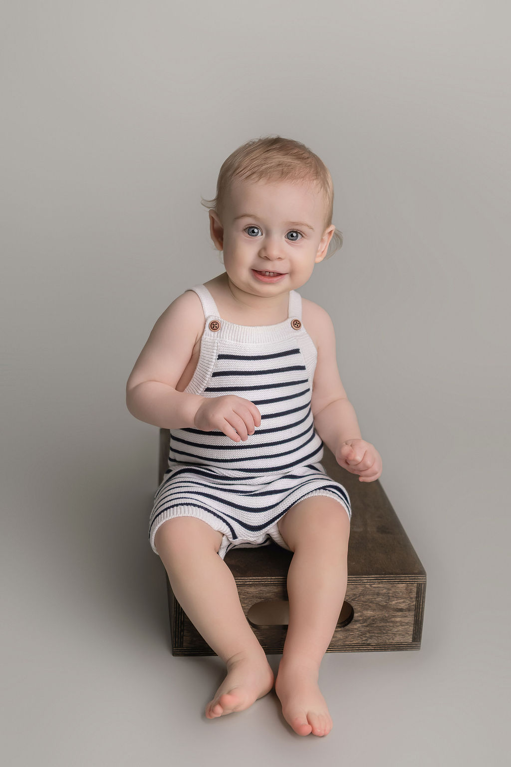 A happy toddler in stripe knit overalls sits on a wooden apple box in a studio before visiting Awesome toy store in stamford ct